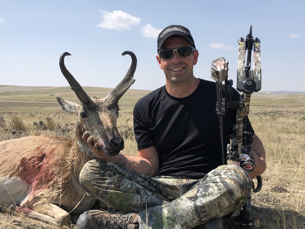Man wearing a black hat and sunglasses holding a bow and sitting next to his trophy antelope
