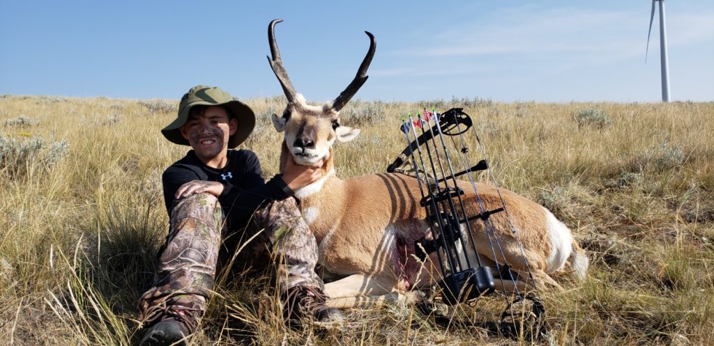 A young boy wearing camo pants and black shirt sitting next to his trophy antelope with a bow leaning against it