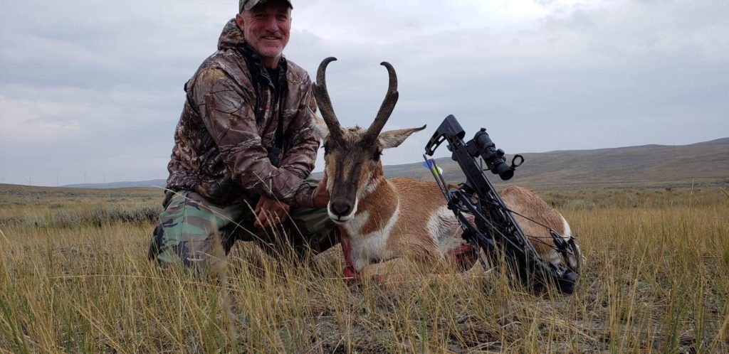 A man wearing full camouflage gear and sunglasses sitting next to and holding his trophy antelope.