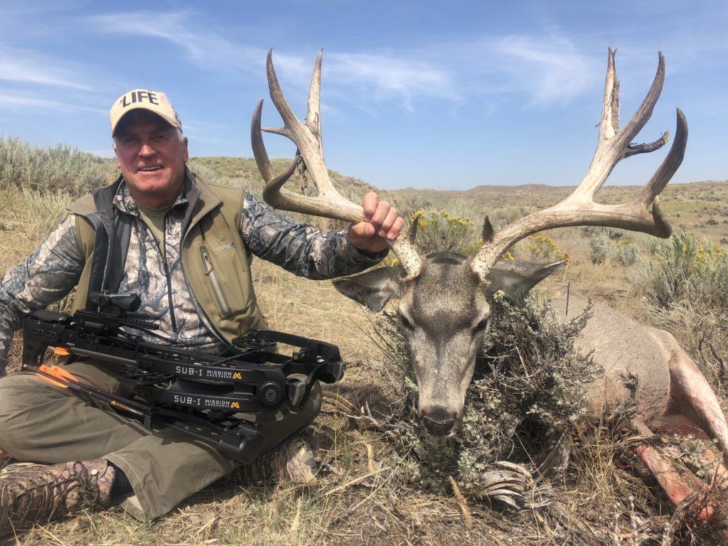 A man with a hat sitting next to his trophy deer after an archery mule deer hunting trip