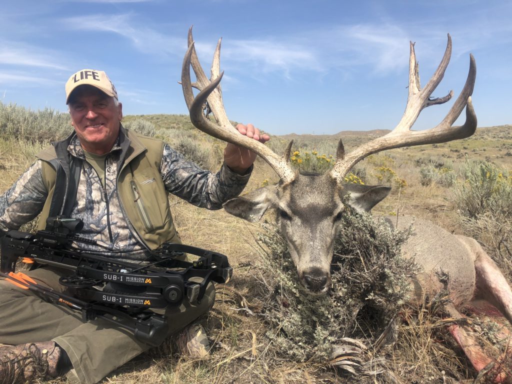 A man with a hat sitting next to his trophy deer after an archery mule deer hunting trip