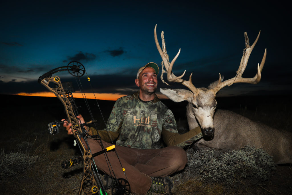 A man holding his trophy deer by the chin with his hunting bow in the other hand after an archery mule deer hunt