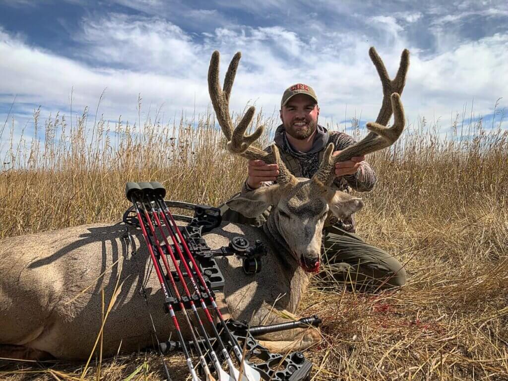 A man holding his trophy deer by the antlers with his hunting bow leaning on it.